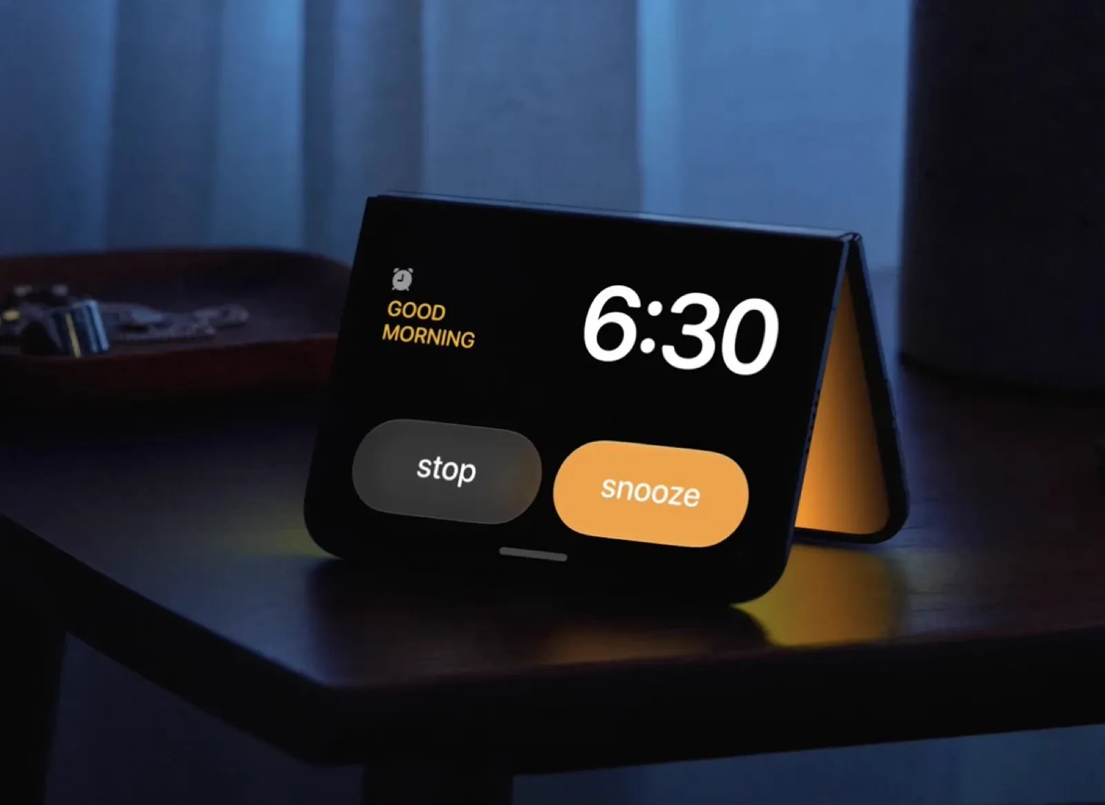 iPhone Duo piegato a tenda sul comodino come sveglia: la scritta Good Morning, l'orario 6:30 e i tasti stop e snooze, con una luce ambientale arancione